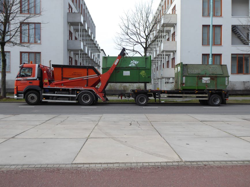 A large flatbed truck equipped with a hydraulic lift arm, parked along a residential street in front of multi-storey apartment buildings with white facades and orange window frames. The truck is carry