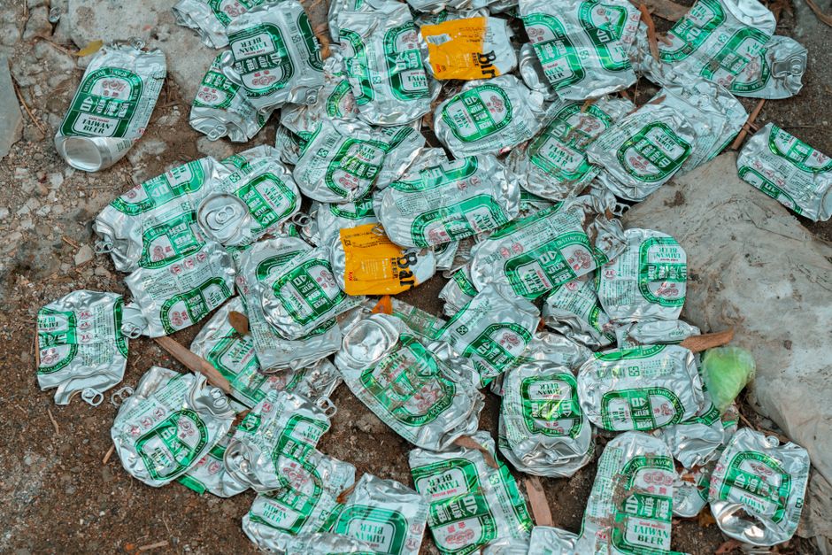The image depicts a collection of overflowing rubbish bins and scattered waste on a paved urban street corner, with various types of refuse including cardboard boxes, paper bags, plastic bottles, and